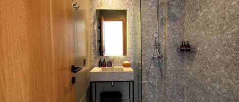 Un baño moderno con puerta de madera, pared de ducha de mármol, ducha con mampara de vidrio y lavamanos con artículos de aseo.