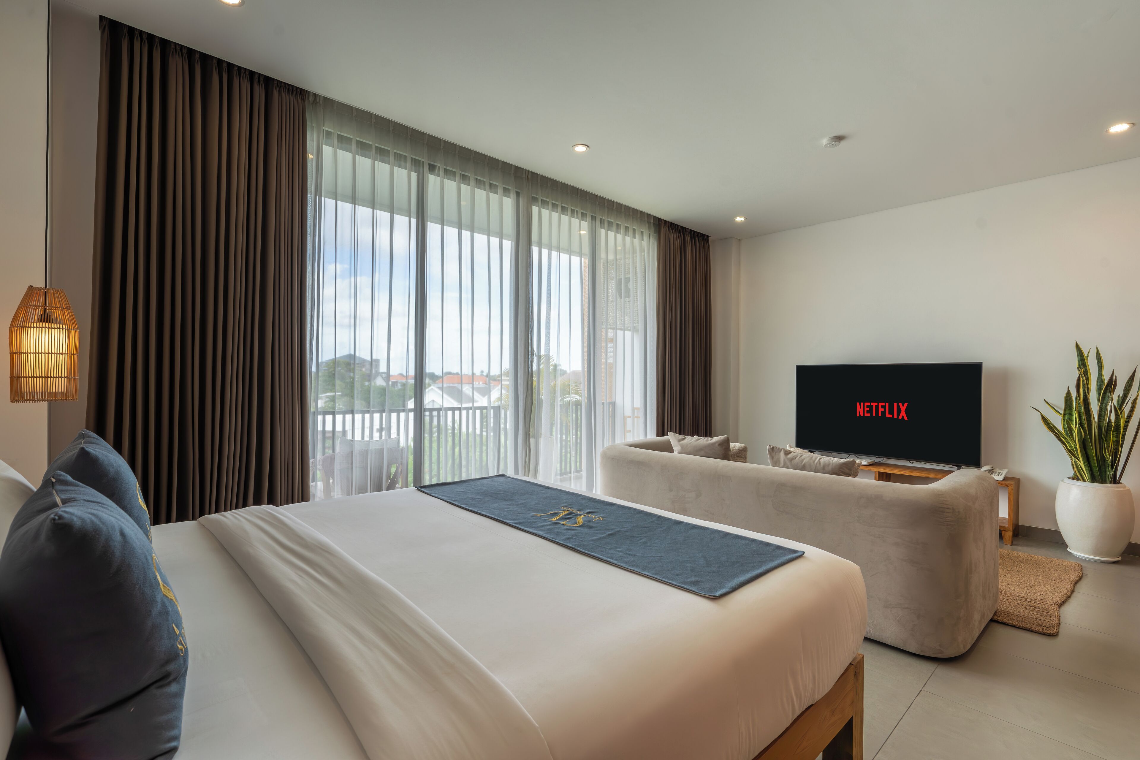 A modern hotel room with a large bed, a sofa, a TV displaying Netflix, and a view of the city.