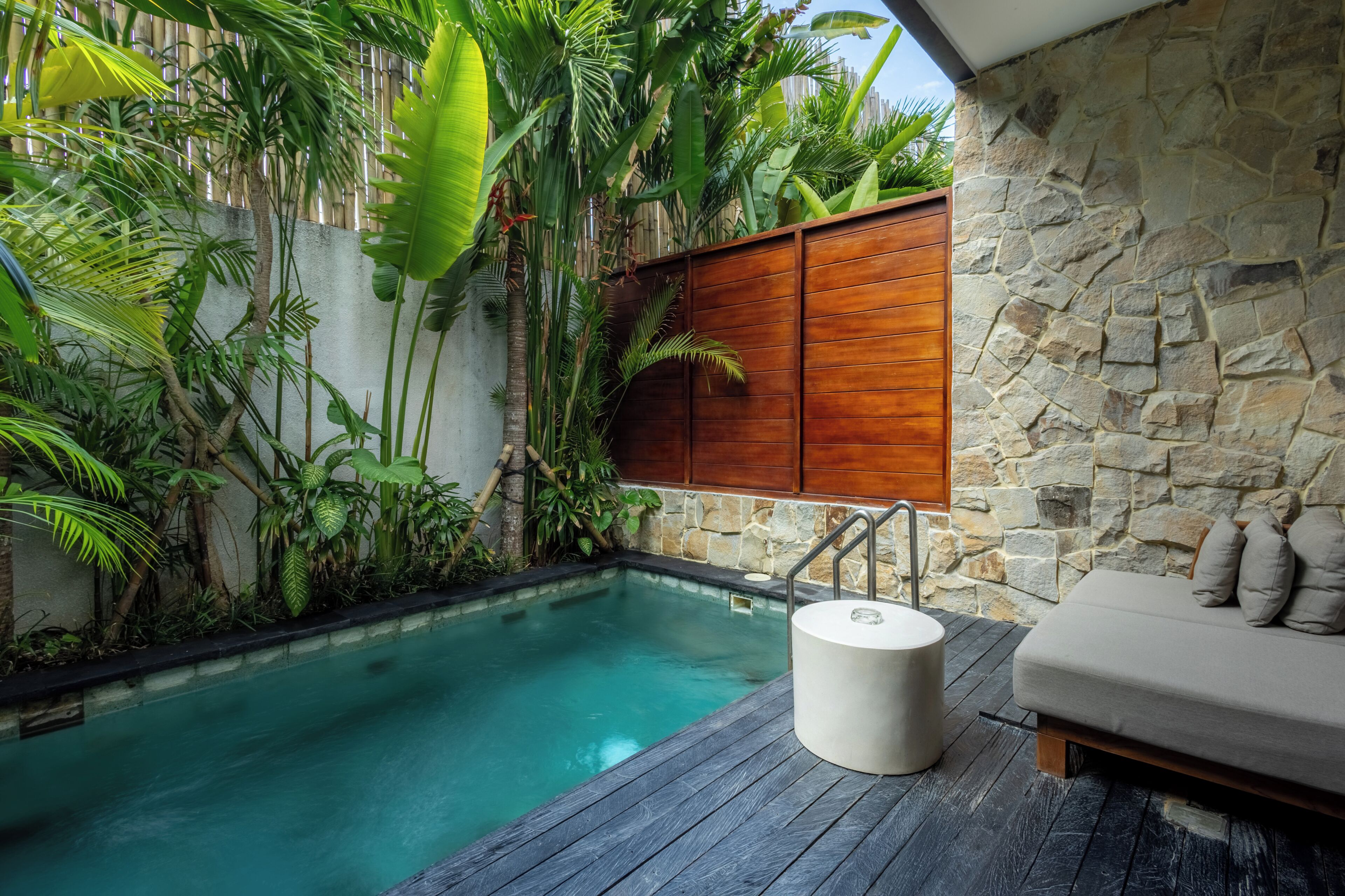 Ein Pool, umgeben von üppiger tropischer Vegetation, einer Steinmauer und einem Holzdeck mit einem weißen Podest.
