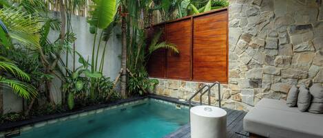 Ein Pool, umgeben von üppiger tropischer Vegetation, einer Steinmauer und einem Holzdeck mit einem weißen Podest.