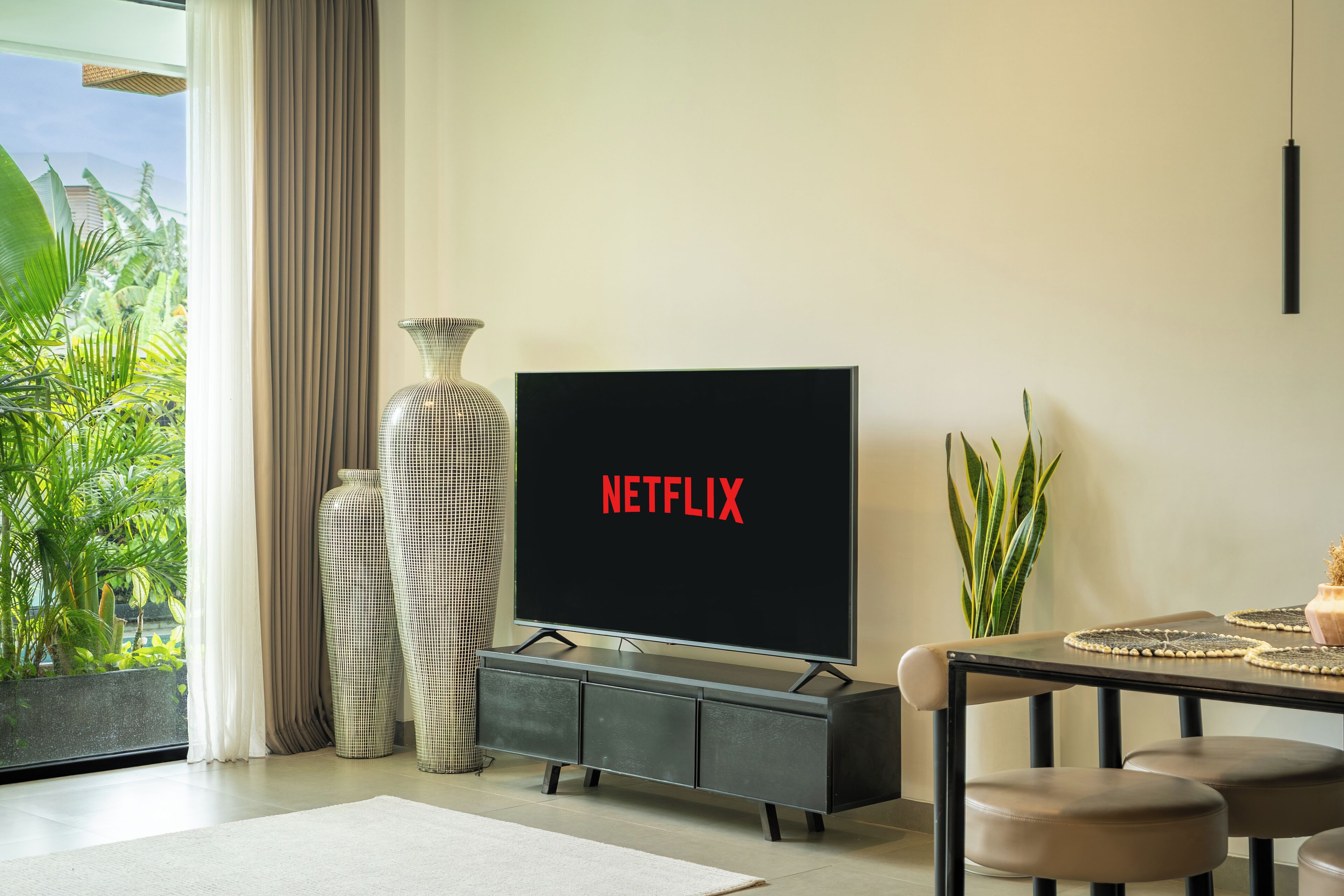 A modern living room with a large flat-screen TV displaying "NETFLIX," a grey entertainment unit, and a dining area with a table and chairs.