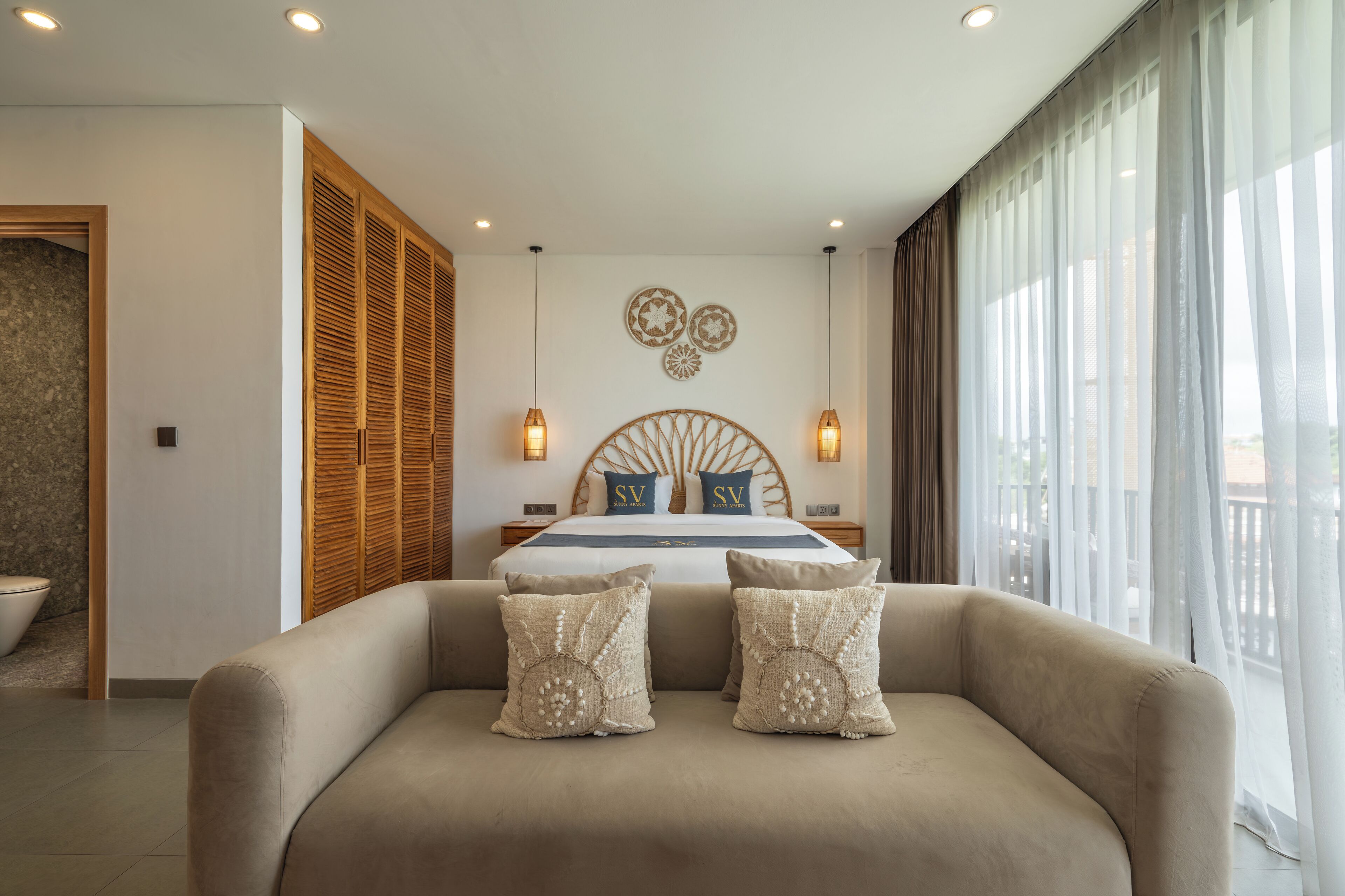 A modern bedroom with a large bed, wooden headboard, two bedside lamps, and a view of the outdoors through sheer curtains.
