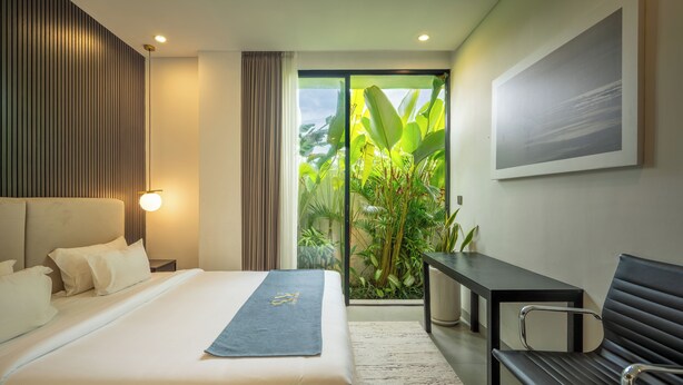 A modern hotel room with a bed, a desk, a bench, and a large window with a view of greenery. - Sunny Aparts 1 by Betterplace (Tibubeneng)
