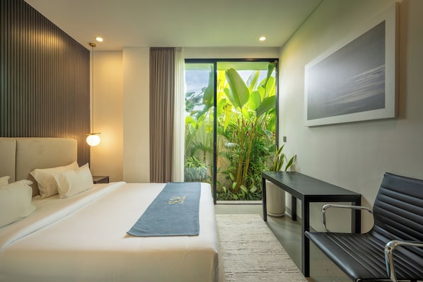 A modern hotel room with a bed, a desk, a bench, and a large window with a view of greenery. - Sunny Aparts 1 by Betterplace (Tibubeneng)