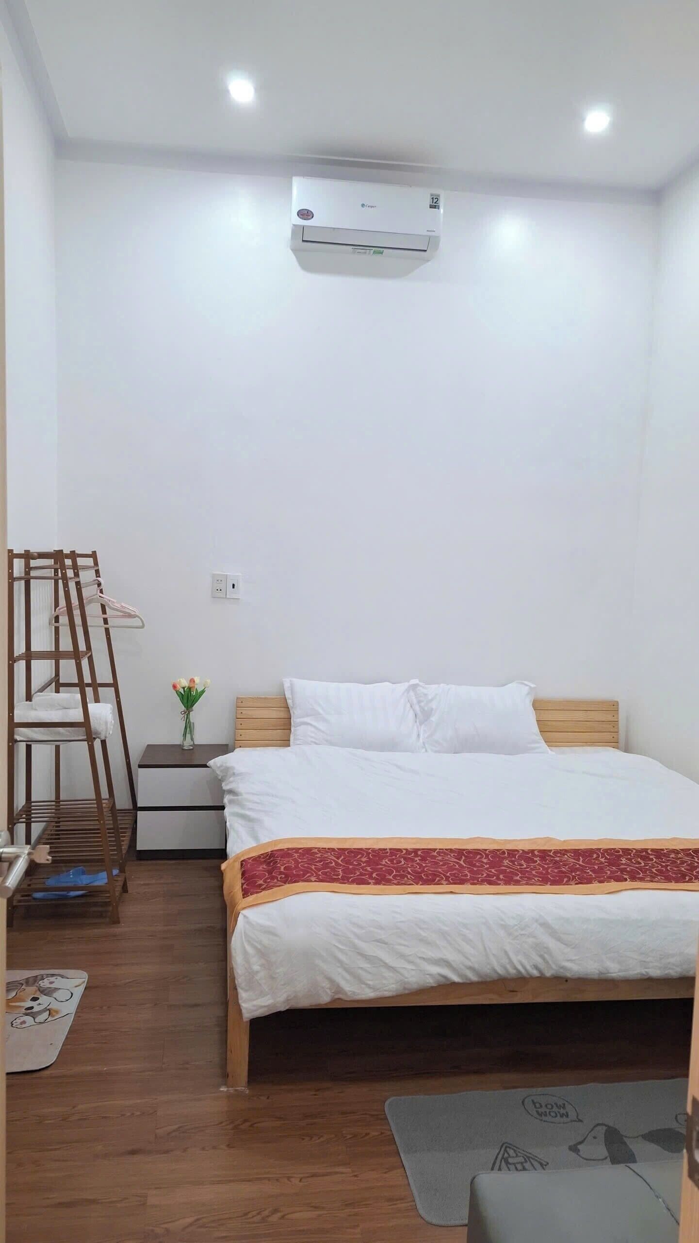 A bedroom with a bed, a wooden nightstand, a wall-mounted air conditioner, a wooden ladder, and a small table with a vase of flowers.