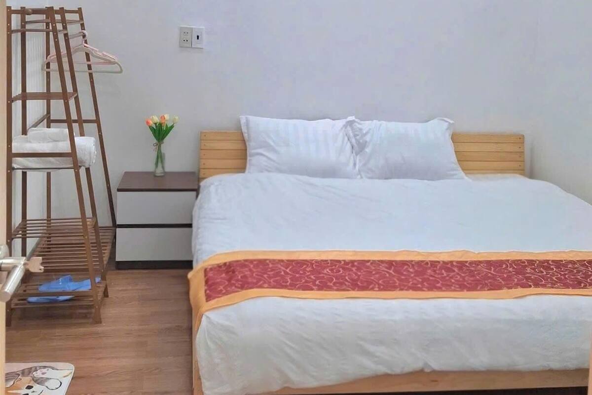 Ένα υπνοδωμάτιο με ένα κρεβάτι, ένα ξύλινο προσκέφαλο, ένα κομοδίνο, ένα κλιματιστικό τοποθετημένο στον τοίχο, μια ξύλινη σκάλα και ένα μικρό βάζο με λουλούδια.