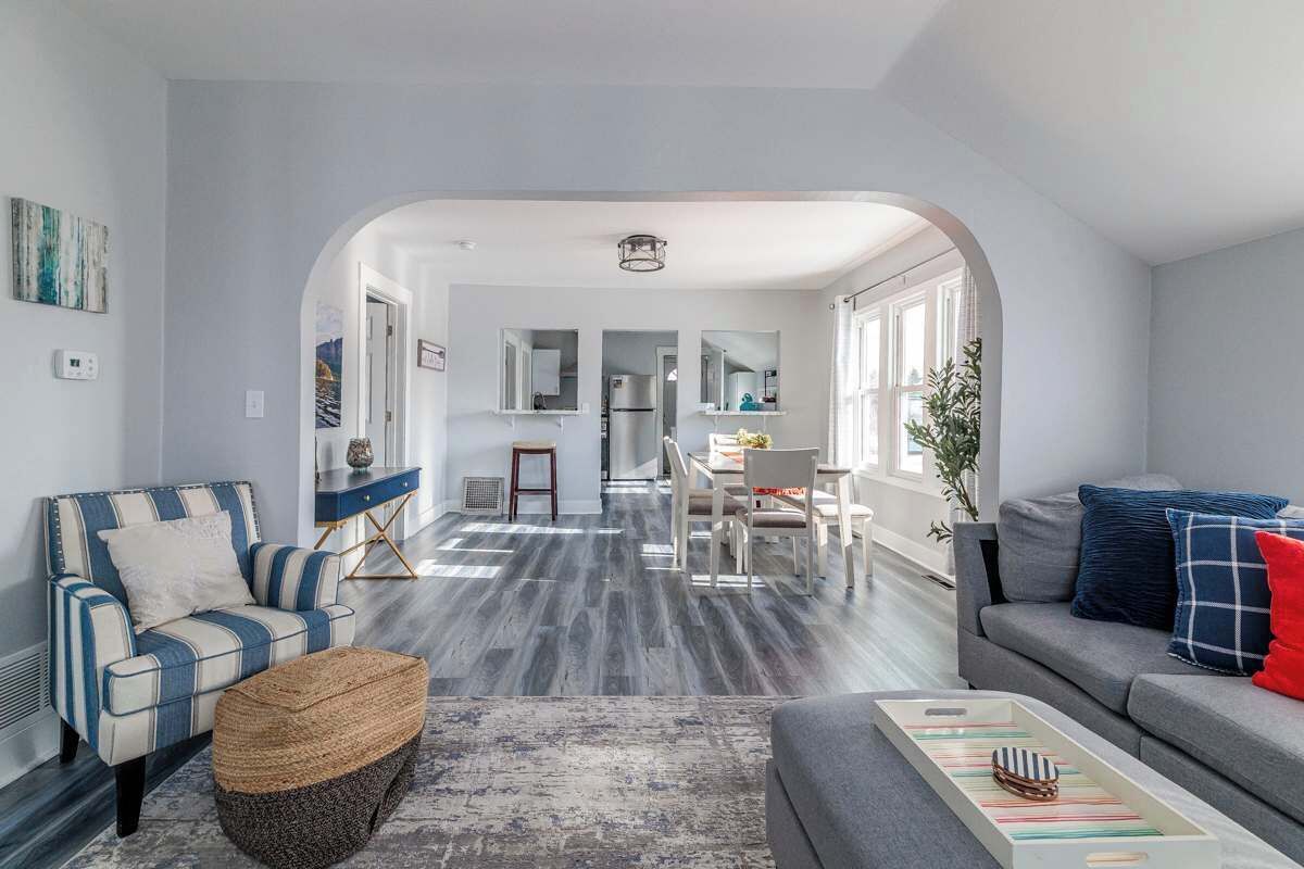 Un salon moderne comprenant un canapé, un fauteuil et un pouf, un coin repas avec une table et des chaises, et une cuisine visible à travers une arche.