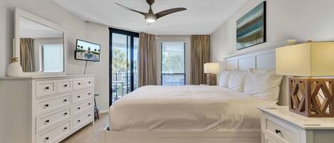 A hotel room with a bed, a TV, a ceiling fan, a desk with a lamp, and a window with curtains.
