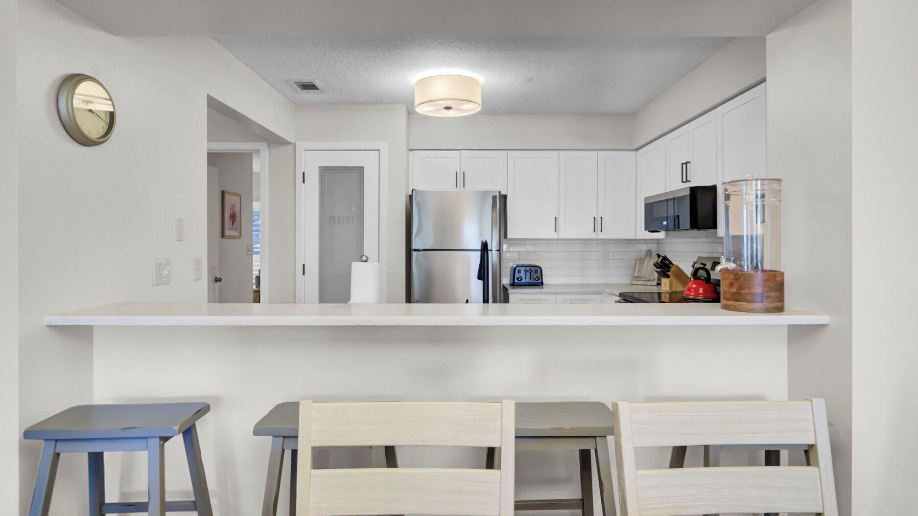 A modern kitchen with white cabinets, a stainless steel refrigerator, a microwave, and a toaster. There are two stools and a table in the foreground.