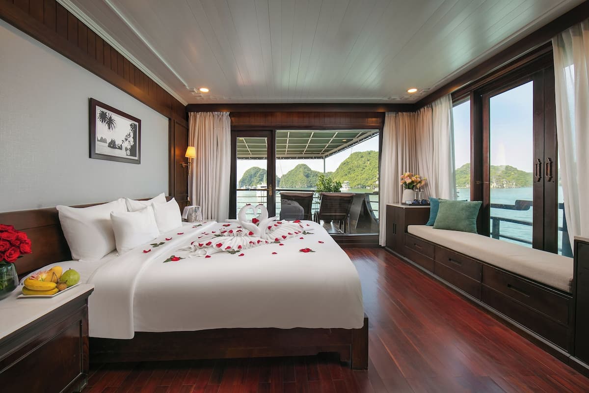 A well-appointed bedroom with a large bed, a bedside table with a fruit bowl, a vase of flowers, and a view of the sea through large windows.