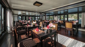 A restaurant with tables set for dining, large windows offering a view, and a ceiling with recessed lighting. - Mila Lux Cruise  (Tuan Chau)