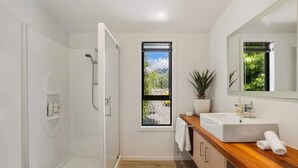 A modern bathroom with a glass shower enclosure, a wooden vanity with a sink, a potted plant, and a window with a view of greenery. - Studio on Martin Lane (Wānaka)