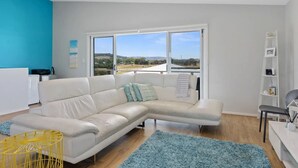 Uma sala de estar moderna com um sofá branco modular, um tapete azul e vista para uma paisagem através de grandes janelas.