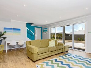 A modern living room with a sofa, a desk, and chairs. There is a large window with a view of the outside. - Splash on Defiance Normanville - Family Fun + Pool (Normanville)