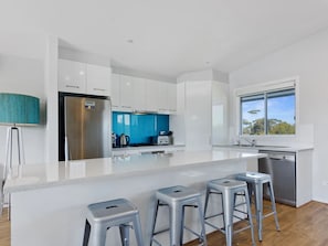 A modern kitchen with white cabinets, a stainless steel refrigerator, a blue backsplash, and a large window. - Splash on Defiance Normanville - Family Fun + Pool (Normanville)