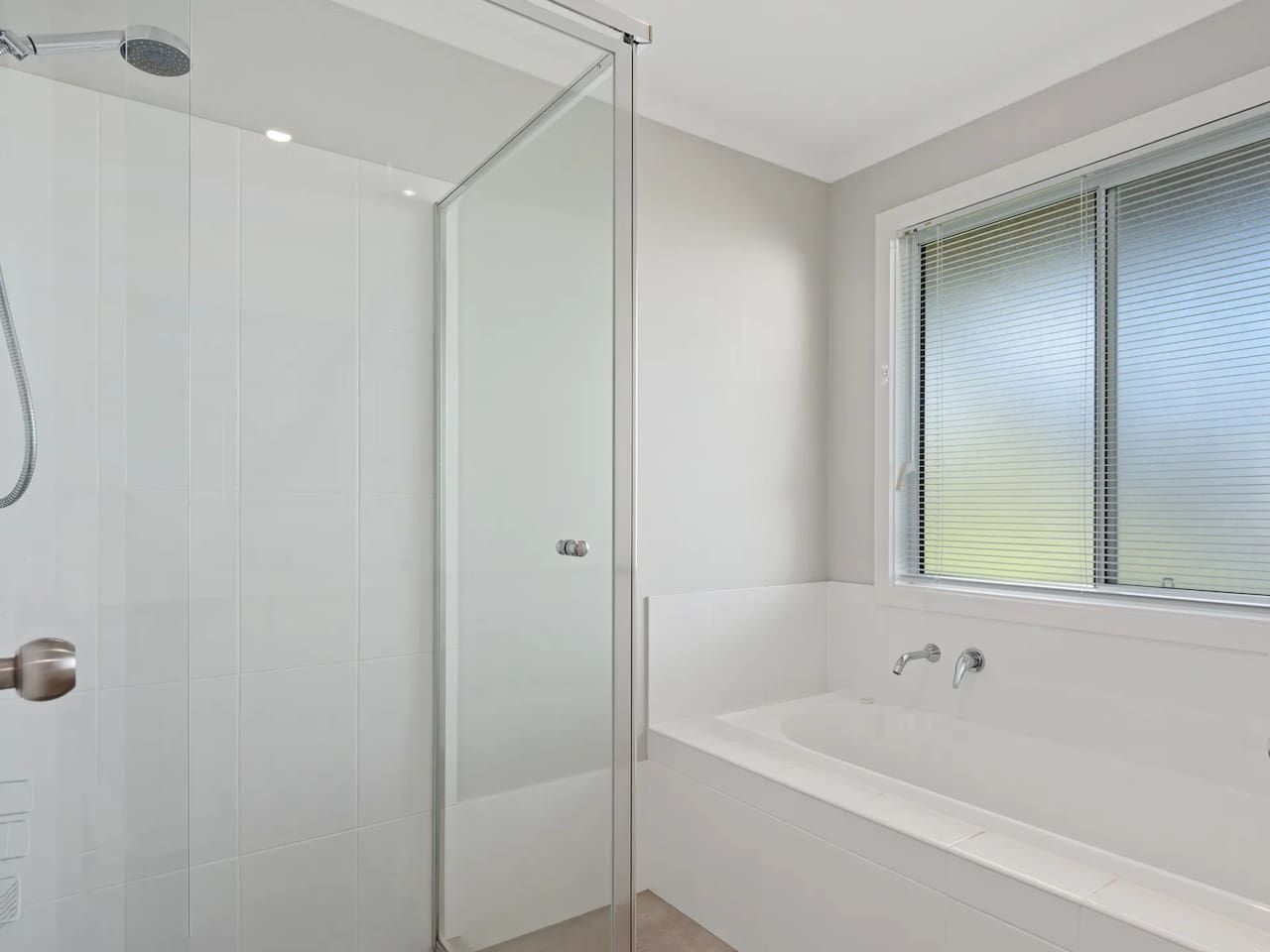 A bathroom with a glass shower enclosure, a bathtub, and a window with blinds.