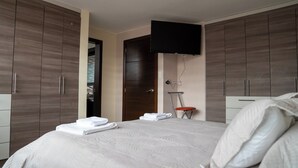 A hotel room with a large bed, wooden wardrobes, a flat-screen TV mounted on the wall, and a small stool. - Vacaciones Seguras - Private Set (Ambato)