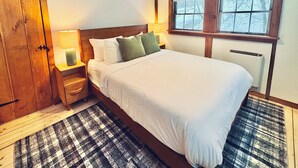 Ein Schlafzimmer mit einem großen Bett, einem Holz-Kopfteil, zwei Nachttischen mit Lampen, einem karierten Teppich und einem Fenster mit Blick auf Bäume.