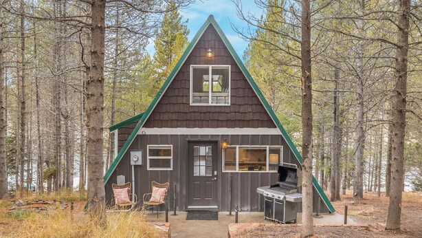 A cabin with a triangular roof, a front door, a small porch with two chairs, and a grill area. - LAKESIDE DOCKSIDE: Henrys Fork Private Beach Dock Fishing Yellowstone UTV Snowmobile Trails (Island Park)