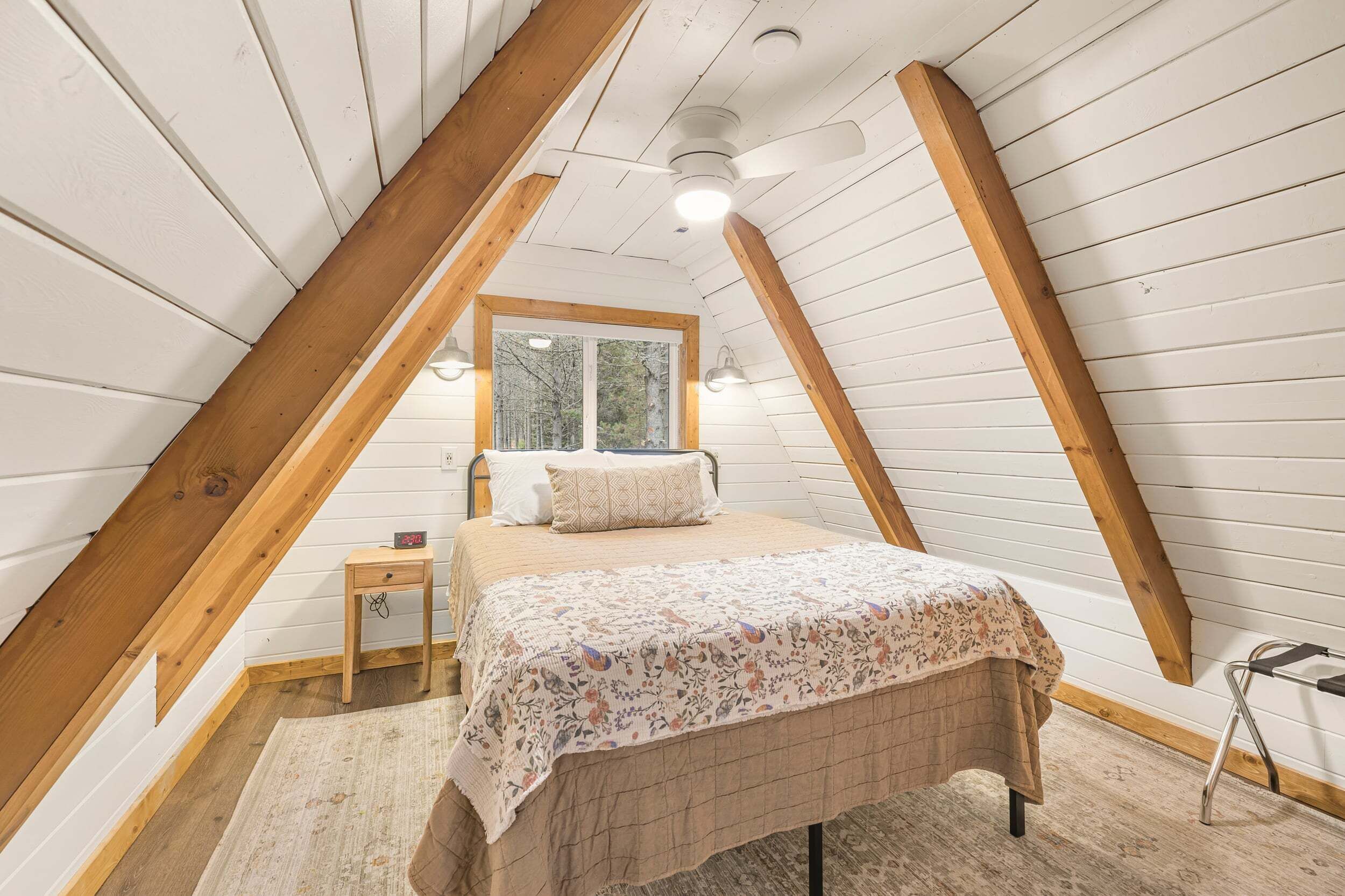 A bedroom with a sloped ceiling, wooden beams, a bed with a floral bedspread, a nightstand, and a window.