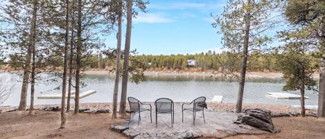 A lakeside seating area with a table and chairs, surrounded by trees and a view of the lake.