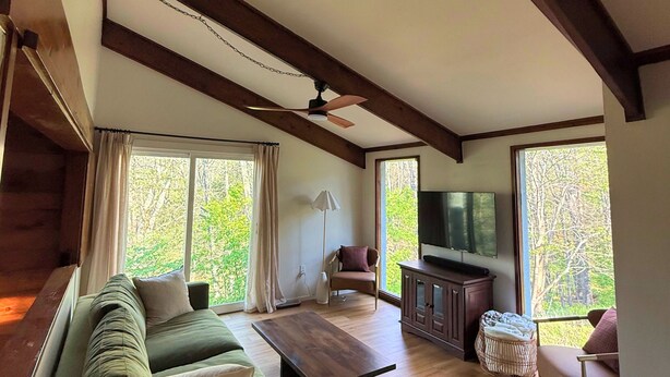 Ein modernes Wohnzimmer mit einer Couch, einem Couchtisch und einem Fernseher, ausgestattet mit großen Fenstern und Holzbalken.