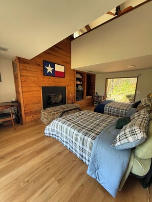 A bedroom with a fireplace, a bed with plaid bedding, a wooden wall with a flag, and a view of trees through a window. - Tranquil bike-in, bike-out chalet in charming Bella Vista (Bella Vista)