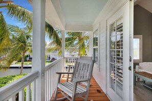 A wooden porch with a rocking chair, a white railing, and a view of a white building with balconies and palm trees. - The Point at Coral Lagoon, Rare End of Canal Villa (Marathon)