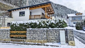 Un chalet moderne doté d’un balcon en bois, entouré d’arbres couverts de neige et d’un mur de pierres.