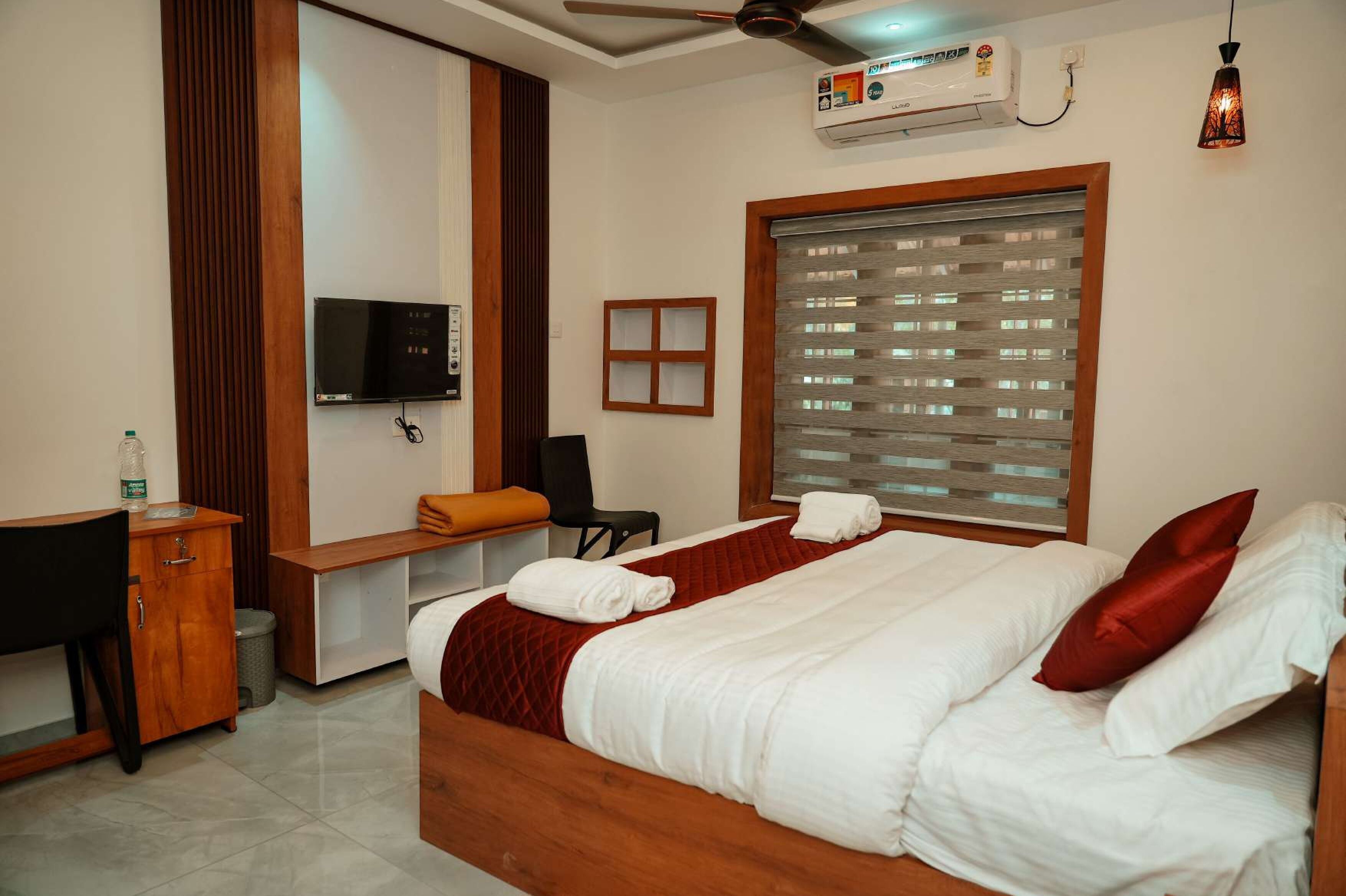 Quarto de hotel com cama, escrivaninha, cadeira, televisão e janela com persianas.