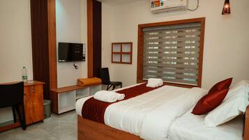 Quarto de hotel com cama, escrivaninha, cadeira, televisão e janela com persianas.