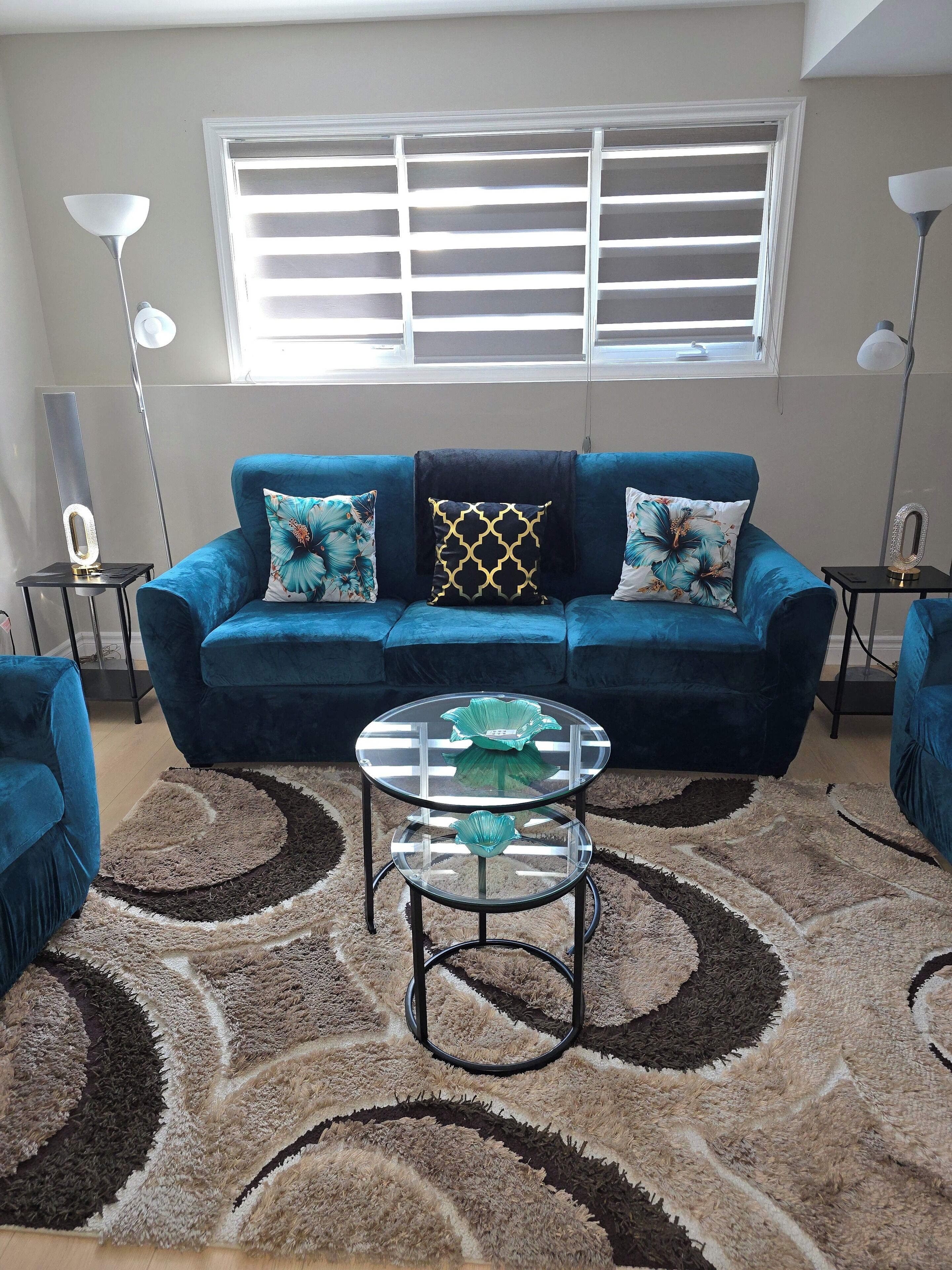 Ein Wohnzimmer mit einem blauen Sofa, zwei Zierkissen, einem Glastisch und einem gemusterten Teppich.