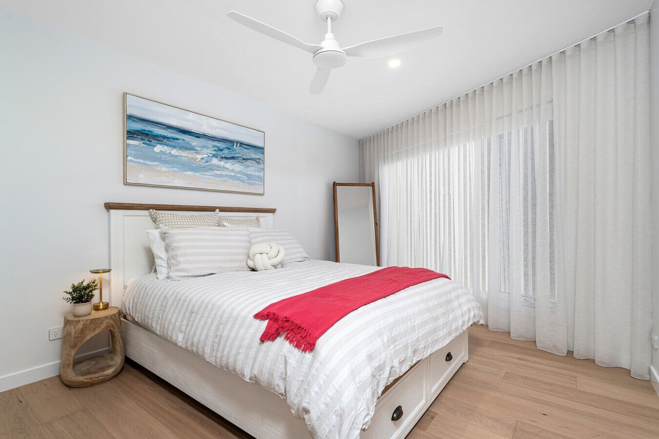 A bedroom with a bed, a ceiling fan, a bedside table with a lamp, a mirror, and a window with curtains.