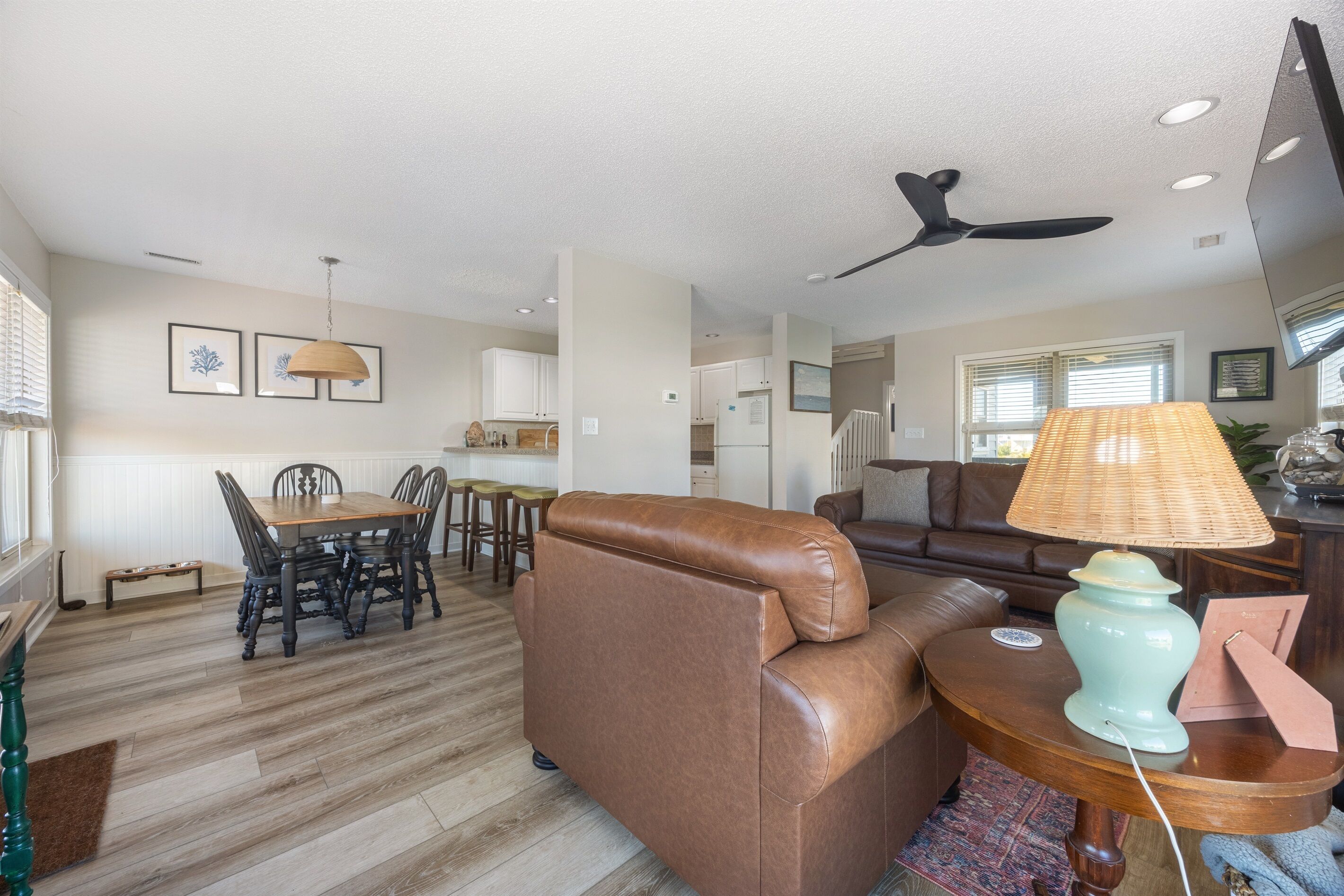 A spacious living room with a brown leather sofa, a dining table with chairs, a ceiling fan, and a lamp on a wooden side table.
