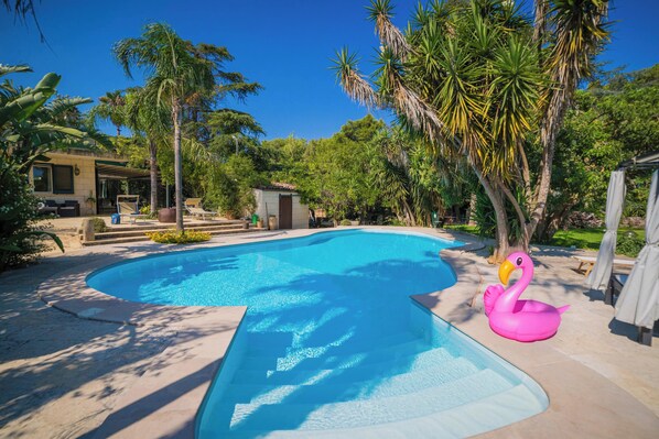 En pool med en rosa uppblåsbar flamingofigur, omgiven av palmer och ett takförsett uteområde.