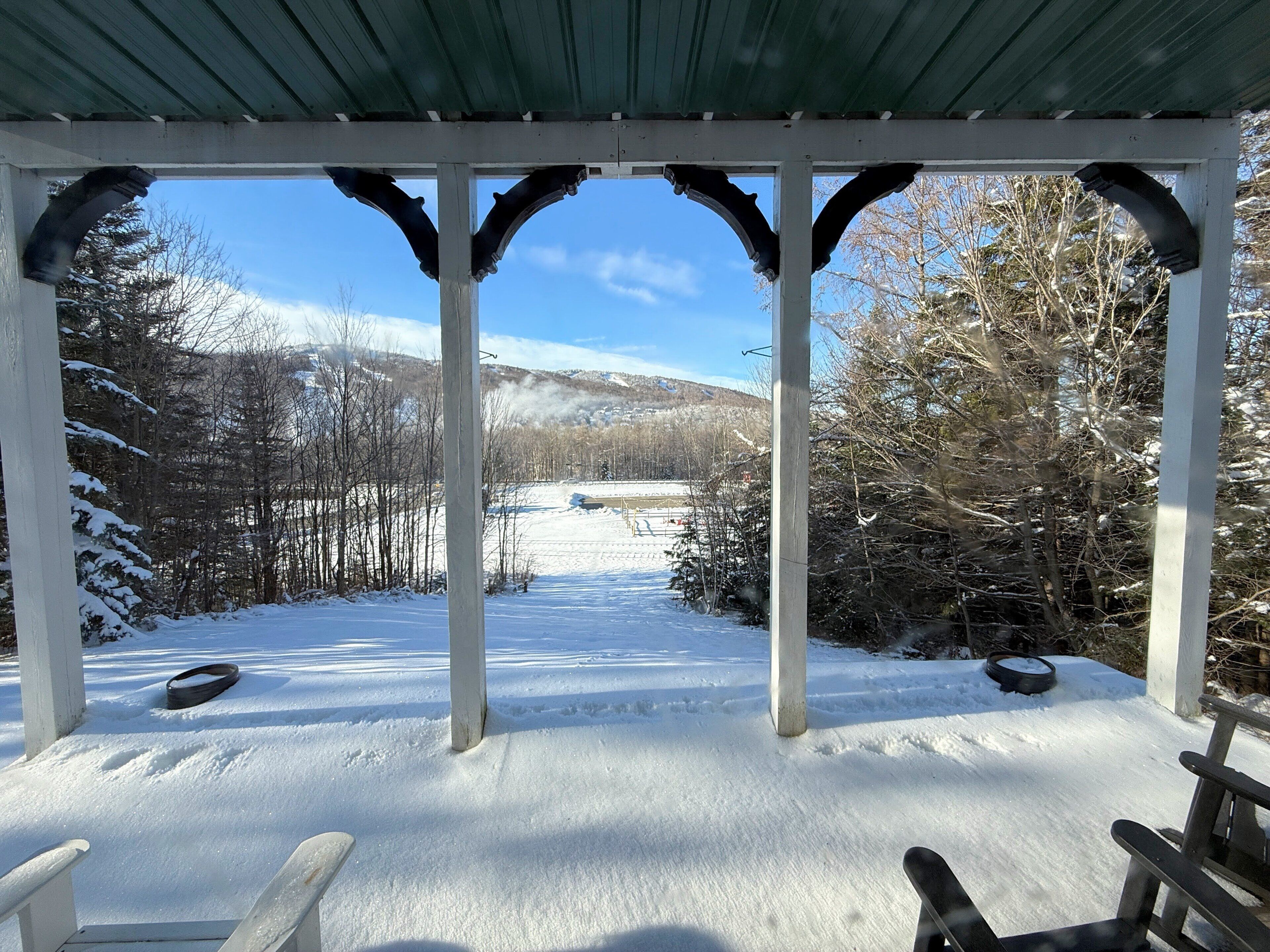 Ein überdachter Außenbereich mit schneebedecktem Boden, Bänken und Blick auf eine verschneite Landschaft mit Bäumen und Hügeln im Hintergrund.