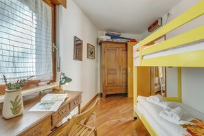 A room with a bunk bed, a desk with a lamp, a window with blinds, and a wooden wardrobe.