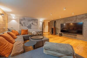 Un salon moderne doté d’un grand téléviseur à écran plat, d’un canapé d’angle avec des coussins orange, d’un pouf rond et d’un plancher en bois.