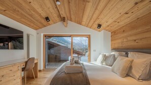 Un dormitorio con techo de madera, una cama grande con almohadas, un escritorio con silla y una puerta corrediza de vidrio que da a un balcón.