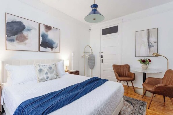 A modern bedroom with a large bed, a bedside table, a chair, a small table with a vase, and abstract wall art. - Chic Studio Apartment on Oglethorpe Square (Savannah)