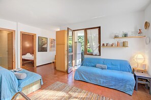 A room with two beds, a wooden wardrobe, a small table with a lamp, and a shelf with decorative items.