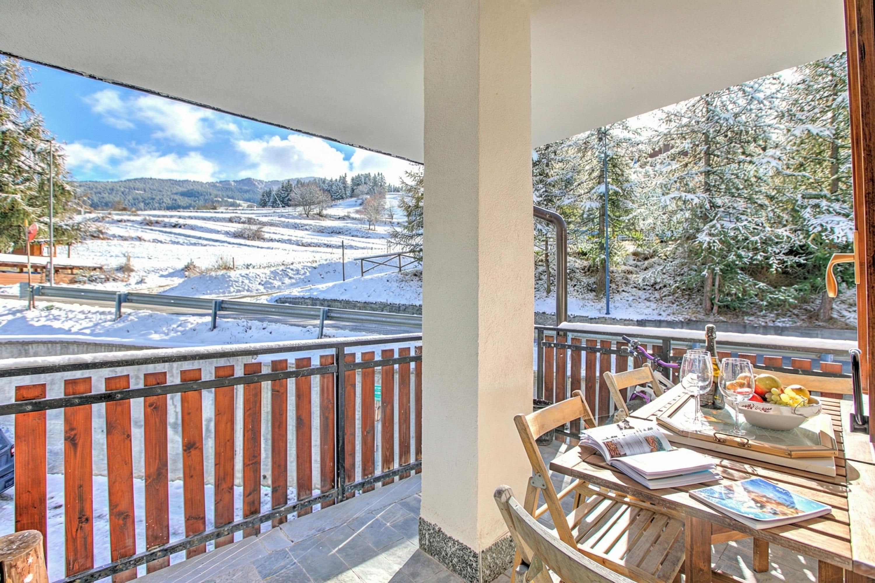 A balcony with a view of a snow-covered landscape, a wooden table with a bowl of fruit, and a bicycle.