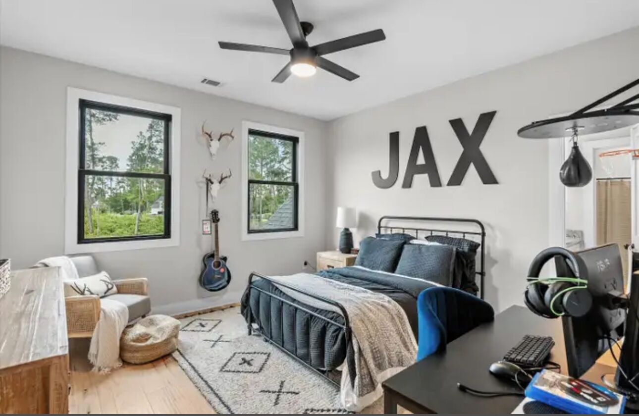 A bedroom with a bed, a desk with a computer, a guitar, and large windows.