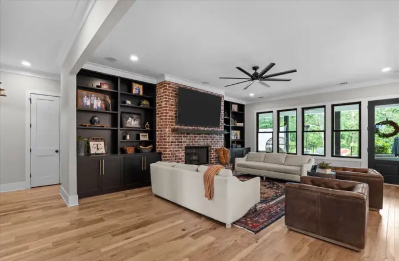 Un salon moderne avec une cheminée en briques, un canapé blanc, un divan en cuir marron et une grande fenêtre donnant sur un paysage verdoyant.