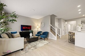 Een moderne woonkamer met een bank, salontafel en stoelen, een televisie waarop 'NETFLIX' staat, en een trap naar een andere kamer.