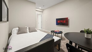 Una camera d'albergo con un letto, un tavolino con una pianta, una TV montata a parete e una porta che dà su un'altra stanza.