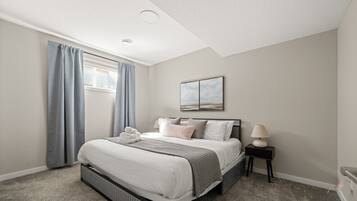A bedroom with a bed, a nightstand, a lamp, a window with curtains, and a painting on the wall.