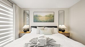 Ein Hotelzimmer mit einem großen Bett, zwei Nachttischlampen, einem Bild an der Wand und einem Fenster mit Jalousien.