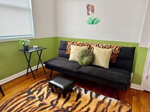 A room with a green accent wall, a black sofa with patterned cushions, a small black table with a plant, and a tiger-striped rug.