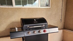 Un barbecue noir pour l’extérieur, doté de quatre brûleurs et d’une étagère latérale, placé contre un mur beige, avec une fenêtre au-dessus.